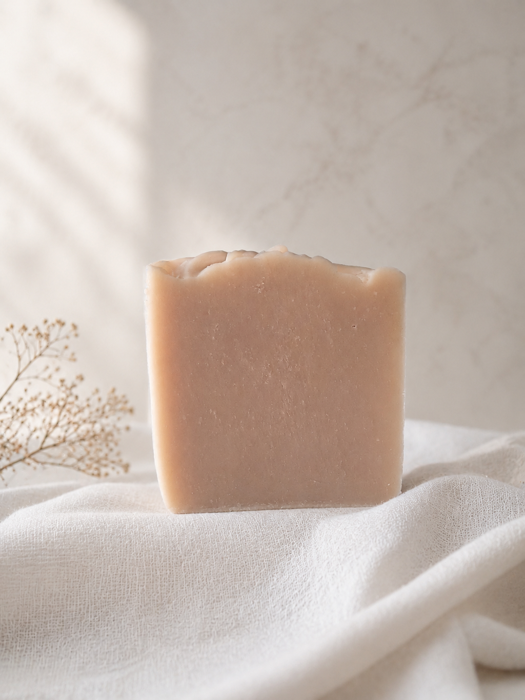 A rectangular bar of pale peach-beige soap standing upright on draped white fabric, with soft natural light and a blurred neutral background.